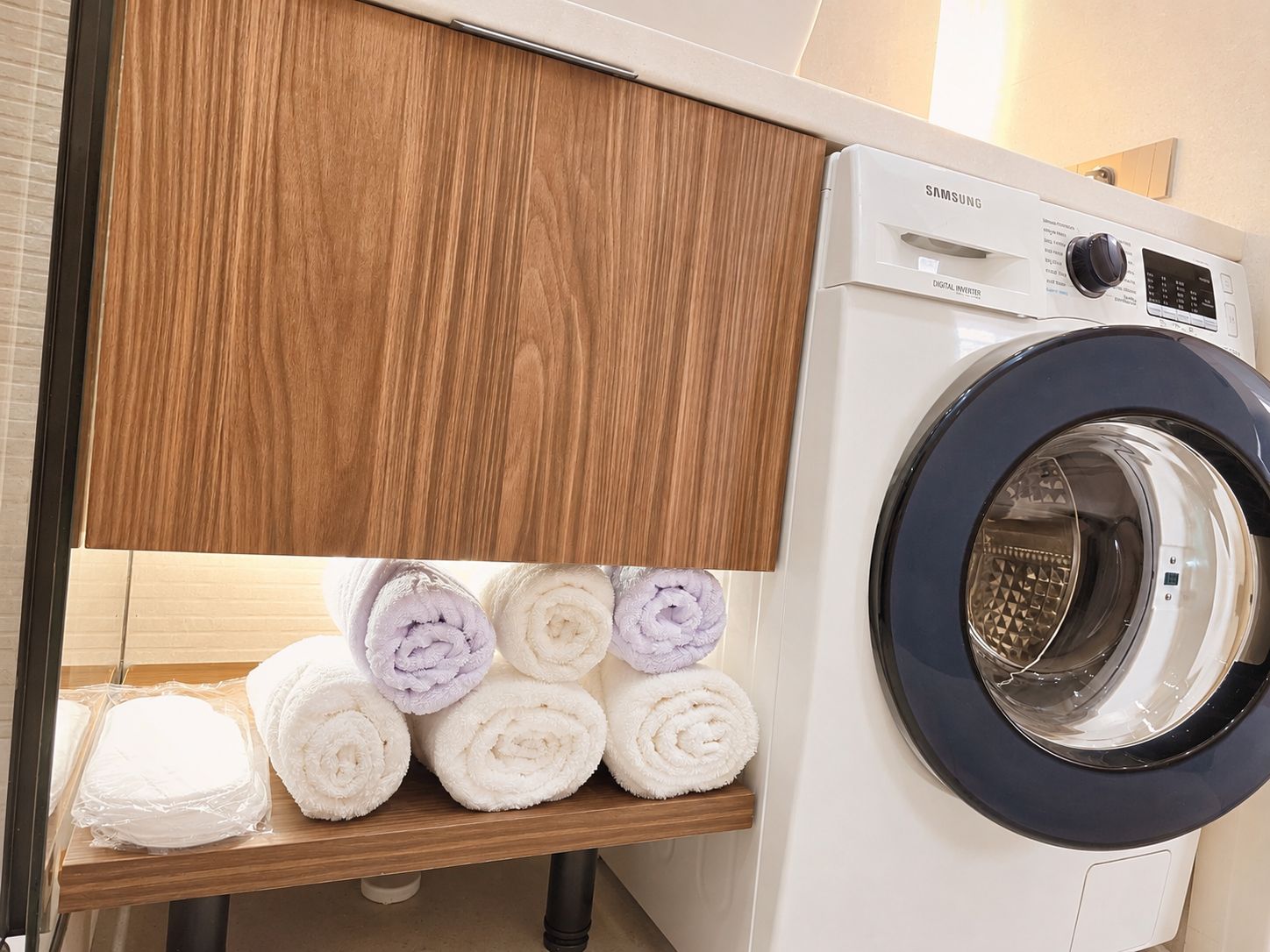 Un lave-linge à variateur de vitesse numérique de marque Samsung, avec une étagère en bois en dessous, sur laquelle sont soigneusement pliées des serviettes.