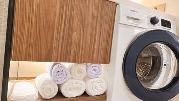 Un lave-linge à variateur de vitesse numérique de marque Samsung, avec une étagère en bois en dessous, sur laquelle sont soigneusement pliées des serviettes.