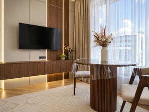 Ein modernes Hotelzimmer mit einem an der Wand befestigten Flachbildfernseher, einem runden Tisch mit einer Vase getrockneter Blumen und einem großen Fenster mit durchsichtigen Vorhängen.