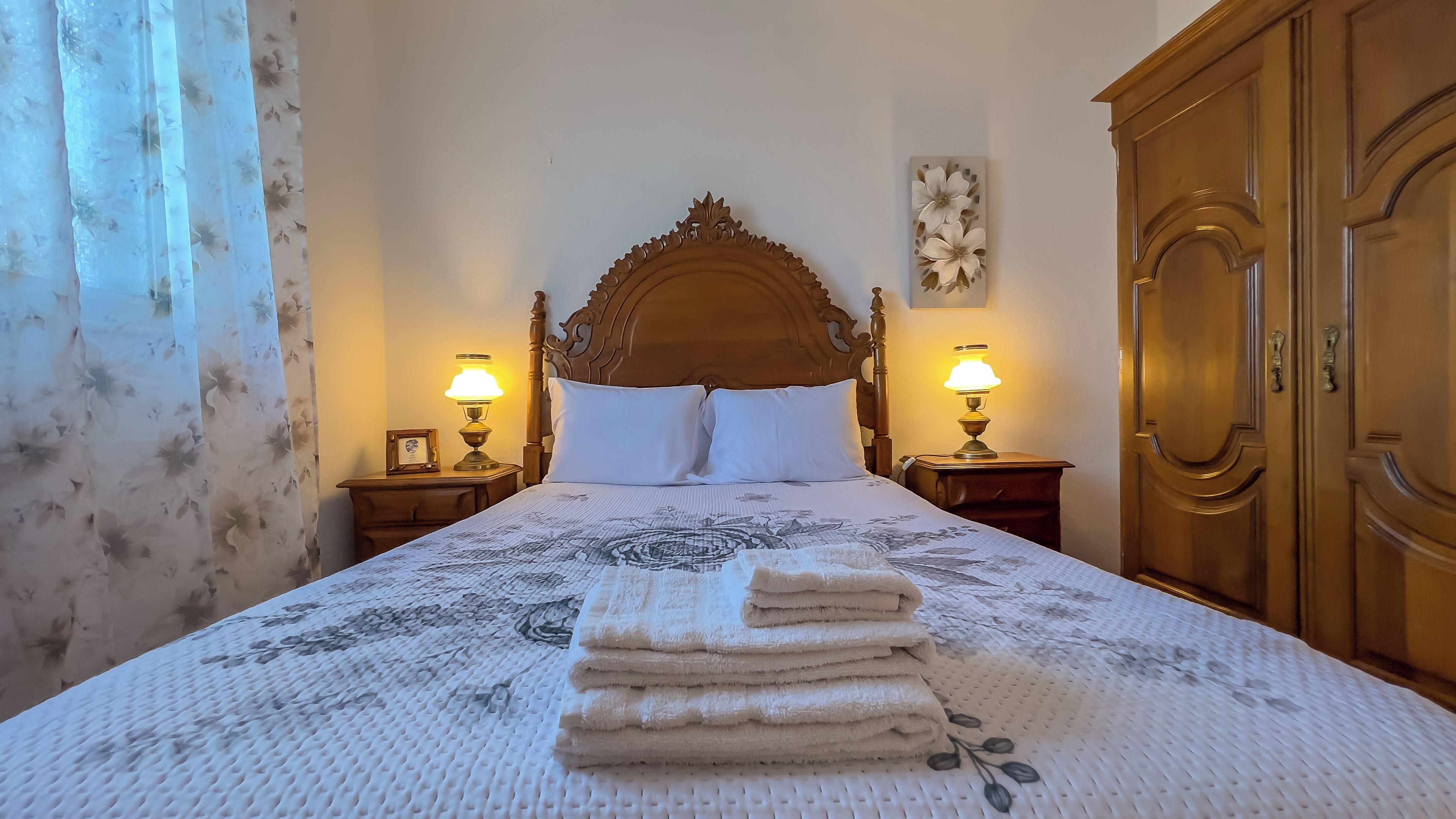 A neatly made bed with a floral-patterned bedspread, two bedside lamps, a wooden headboard, and a wooden wardrobe.