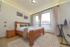 A bedroom with a wooden bed, a bedside table, a lamp, a window with curtains, and a view of buildings outside. - Cozy&stlyish  1-bedroom apartment in Riara One Residency Lavington Nairobi (Nairobi)