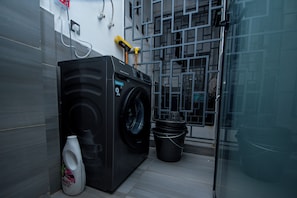 A compact laundry room with a front-loading washing machine, a bucket, and a bottle of Ariel Power Gel. - Cozy&stlyish  1-bedroom apartment in Riara One Residency Lavington Nairobi (Nairobi)
