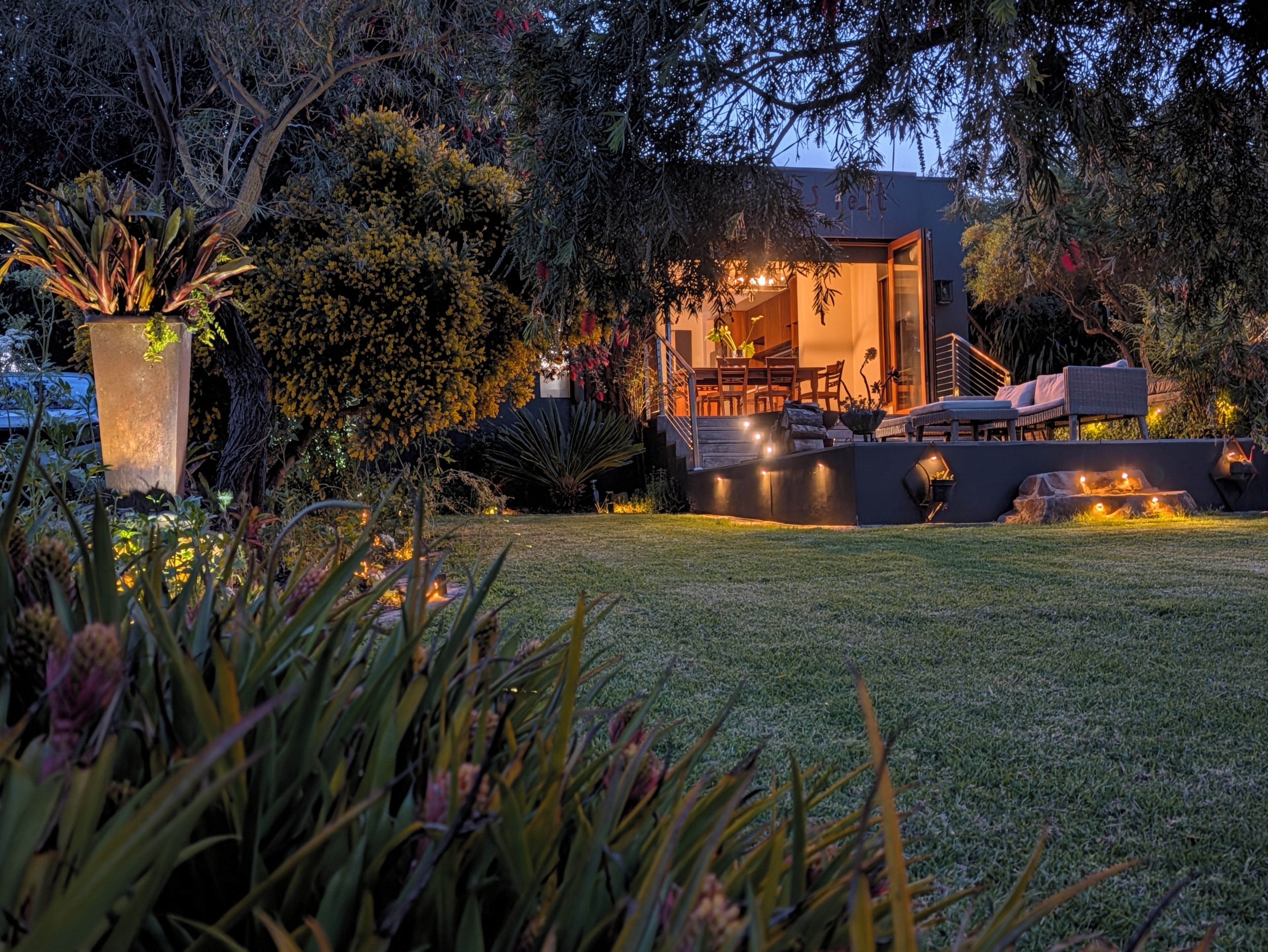 Un área de estar moderna al aire libre con piscina, rodeada de exuberante vegetación y una entrada bien iluminada.