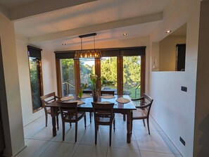 A dining area with a table set for four, surrounded by chairs, a hanging light fixture, and a view of greenery through large windows.