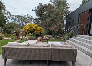 A wicker sofa set on a wooden deck with a fire pit in the center, surrounded by greenery and trees.
