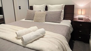 A neatly made bed with a white comforter and two rolled towels, a bedside table with a lamp, and a dark wooden headboard.