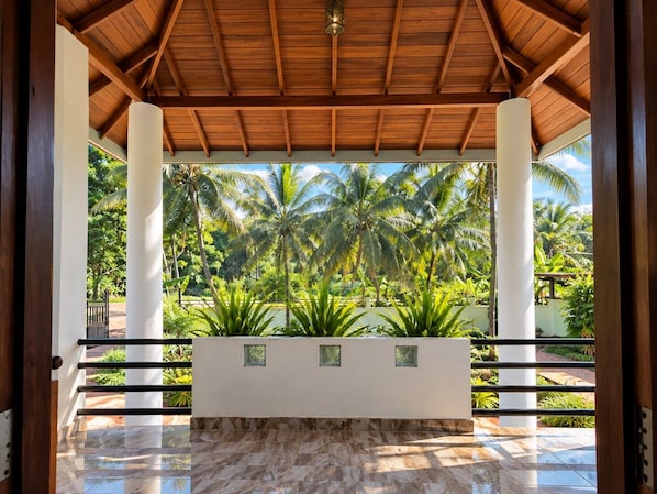 A covered outdoor area with a wooden ceiling, a marble floor, and a view of palm trees.