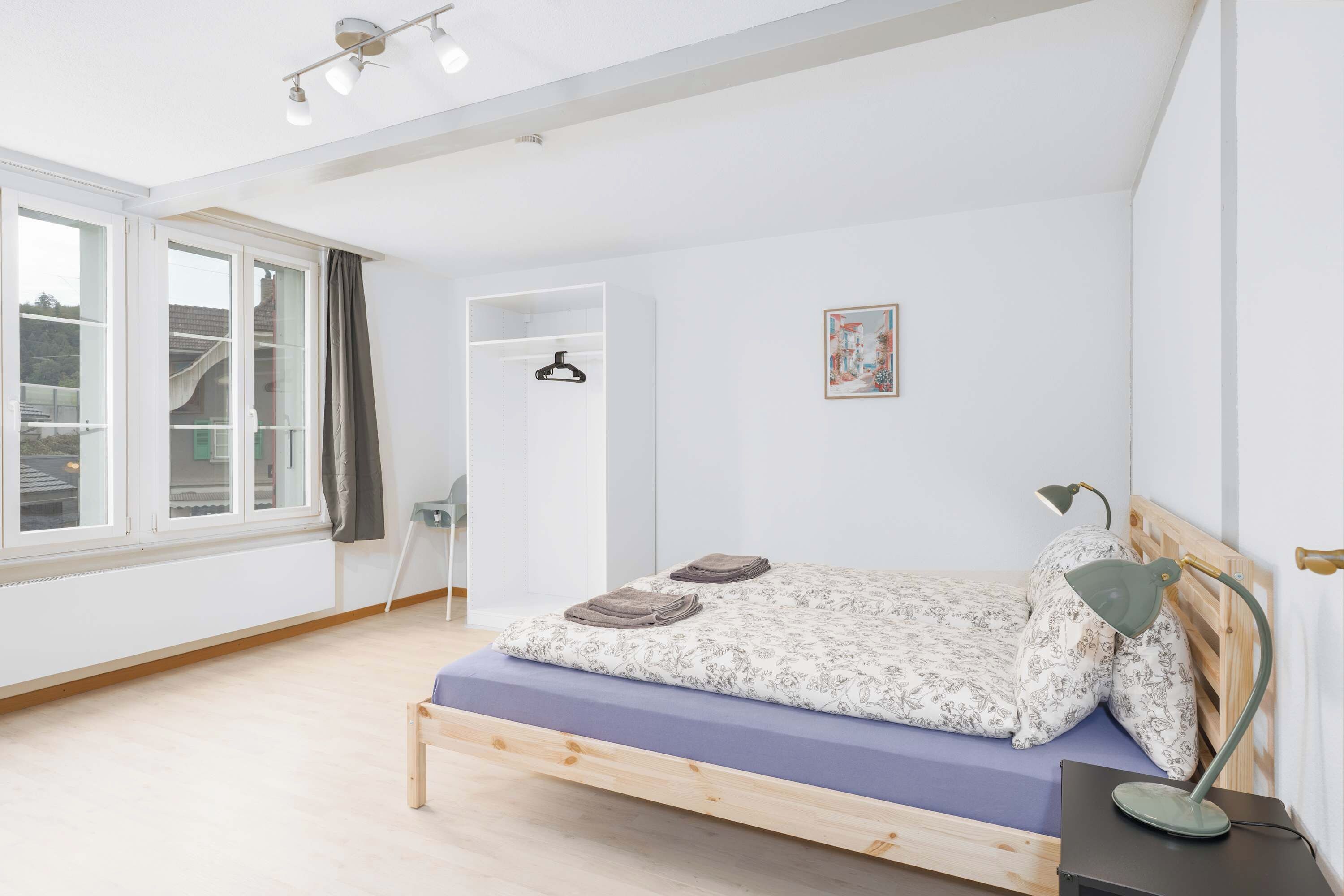 A bedroom with a bed, a bedside table, a lamp, a hanging coat rack, a window with curtains, and a door.