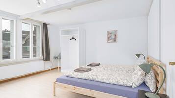 A bedroom with a bed, a bedside table, a lamp, a hanging coat rack, a window with curtains, and a door.
