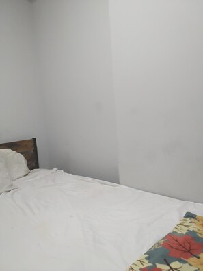 Cama com roupa de cama branca e cabeceira de madeira em quarto com paredes brancas e simples.