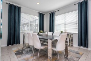 A dining area with a table and chairs, large windows with curtains, and a ceiling light. - Bella Vida 5BR Pool Home with Game Room & Loft (Kissimmee)