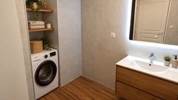 A compact bathroom with a washing machine, wooden shelves, a mirror, and a sink.