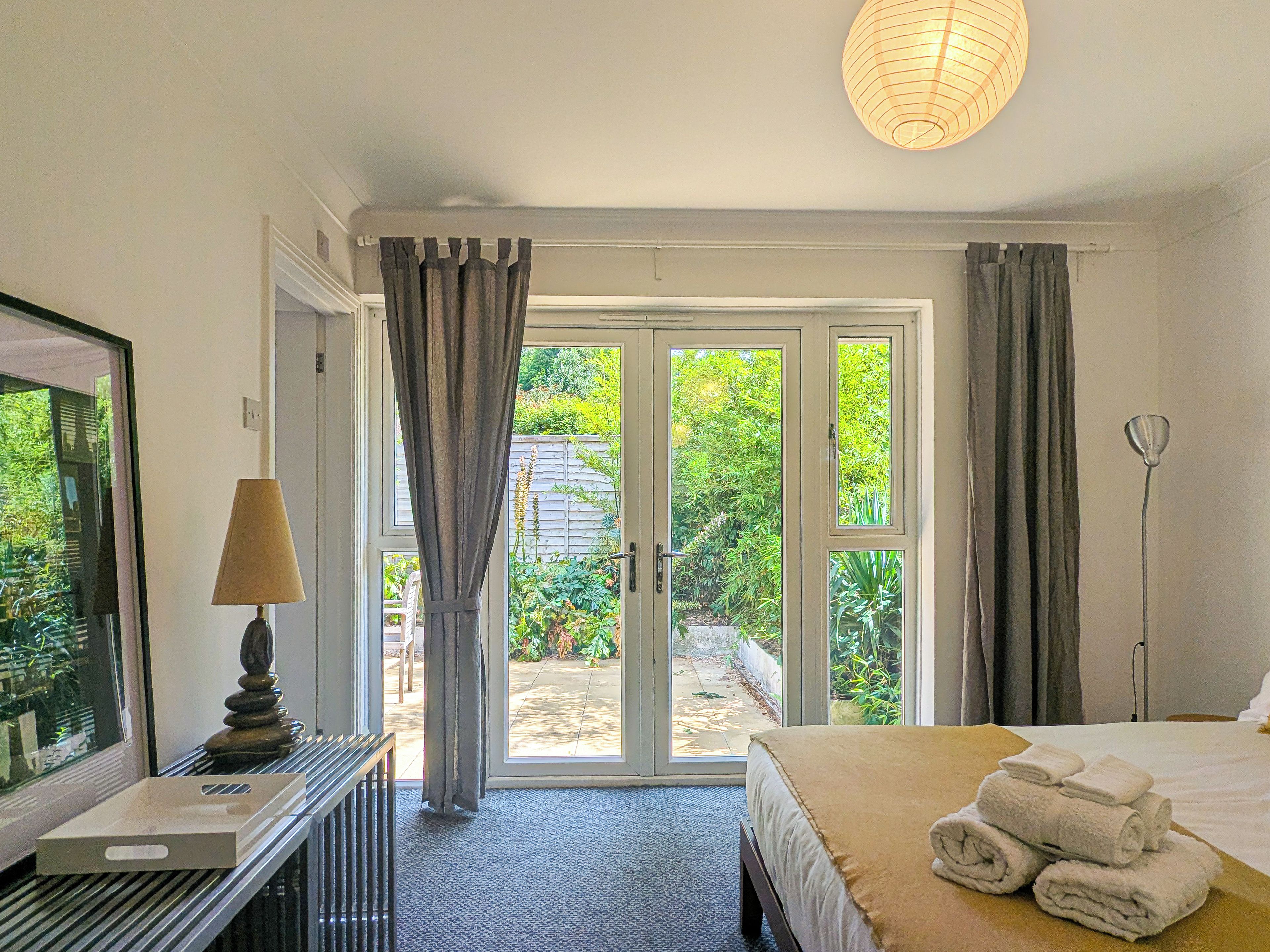A hotel room with a bed, a desk with a lamp, a television, and a view of a garden through the door.