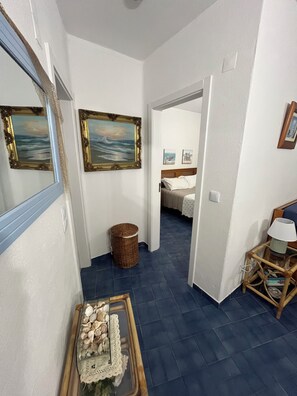 A hallway with a blue-tiled floor, a wicker basket, a glass table with seashells, and framed artwork on the walls. - Edifício Corte Real, Albufeira (Albufeira)
