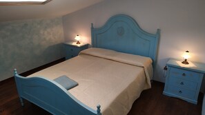 A bedroom with a blue bed, two bedside tables with lamps, a nightstand, and a window with a view. - Mediterranean Idyll (Sardegna)