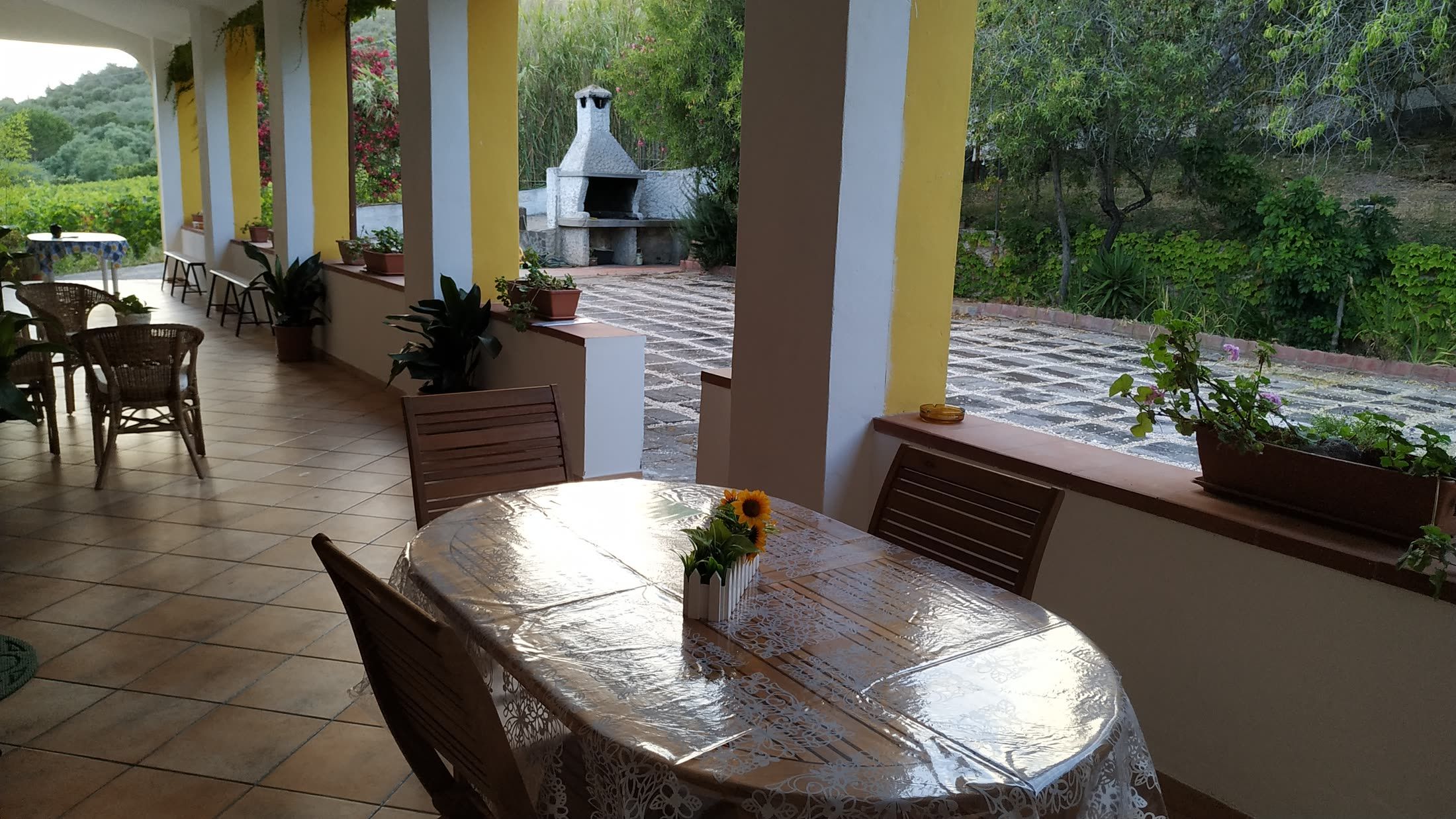 Ein überdachter Patio-Bereich mit Tisch und Stühlen, Blumentöpfen und Blick auf einen Garten.