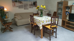 A dining area with a table set for four, surrounded by wooden chairs with yellow cushions. A vase with sunflowers is on the table. - Mediterranean Idyll (Sardegna)