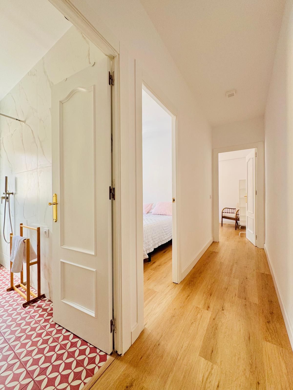A hallway with a wooden floor leading to a bedroom with a bed and a chair.