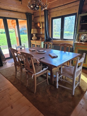 Une table à manger en bois, avec des chaises, un lustre et une vue sur l’extérieur grâce à de grandes fenêtres.