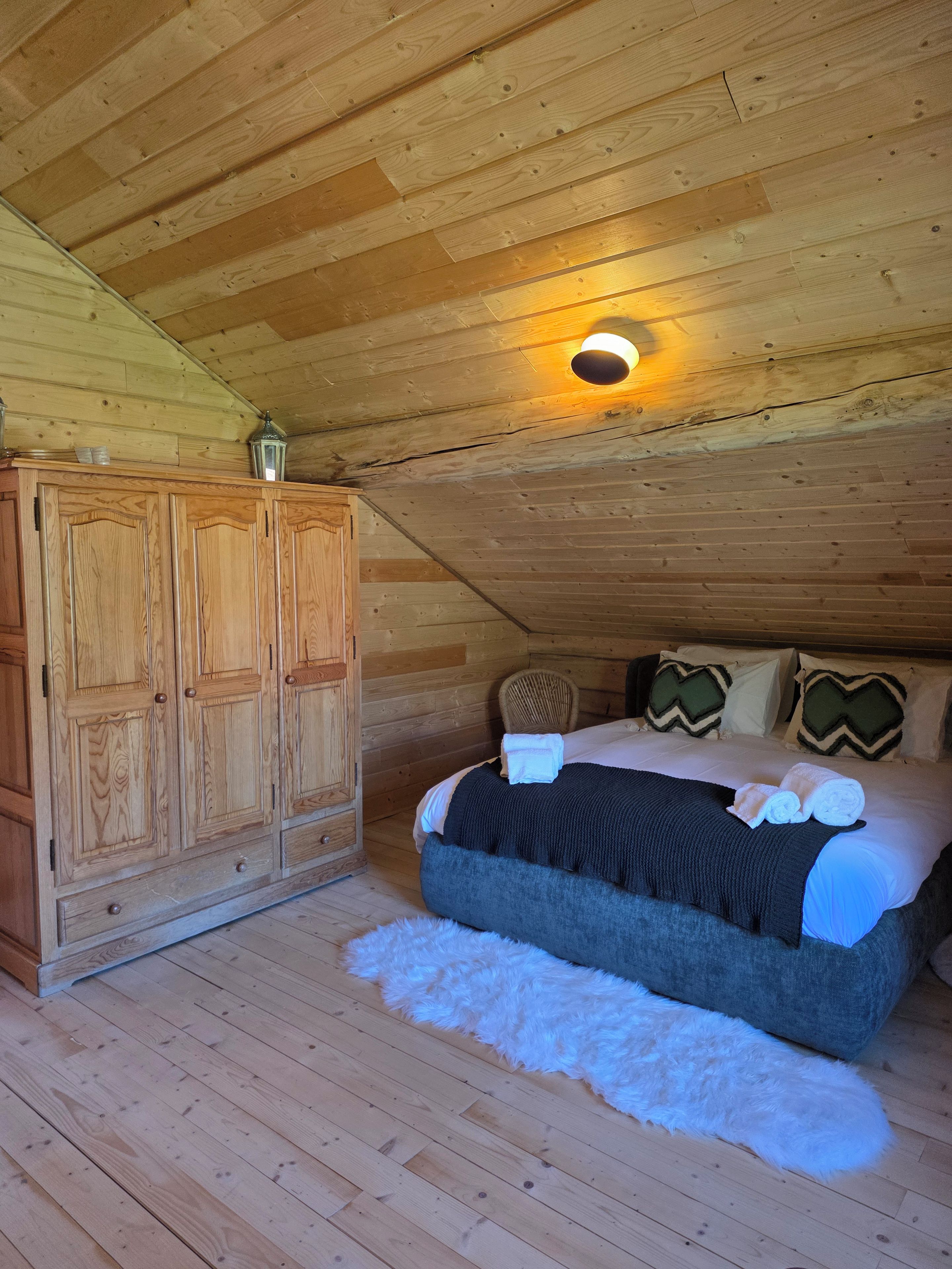 A wooden cabin bedroom with a bed, pillows, a wardrobe, and a small rug.