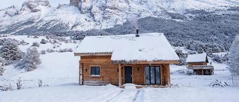 En stuga av trä med ett snötäckt tak, en skorsten och en fri utsikt över snöklädda berg i bakgrunden.