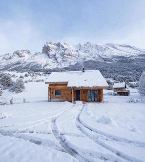 Une cabane en bois, dont le toit est recouvert de neige, avec une cheminée, et qui offre une vue dégagée sur les montagnes enneigées en arrière-plan.