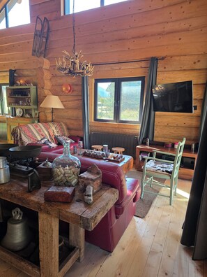 Interior rústico de cabana com mesa de jantar de madeira, sofá vermelho e televisão na parede.