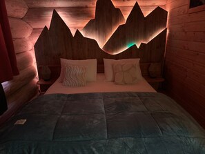 Una habitación con cama, almohadas, una mesita de noche y un diseño de montaña de madera en la pared.