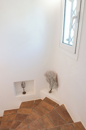 Esquina de una habitación con paredes blancas, una ventana con marco blanco, un estante pequeño con un candelabro de metal y una planta decorativa.