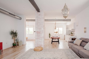 Amplia sala de estar con un pilar de ladrillo, una zona de comedor con una mesa preparada para dos personas, y una cocina al fondo.