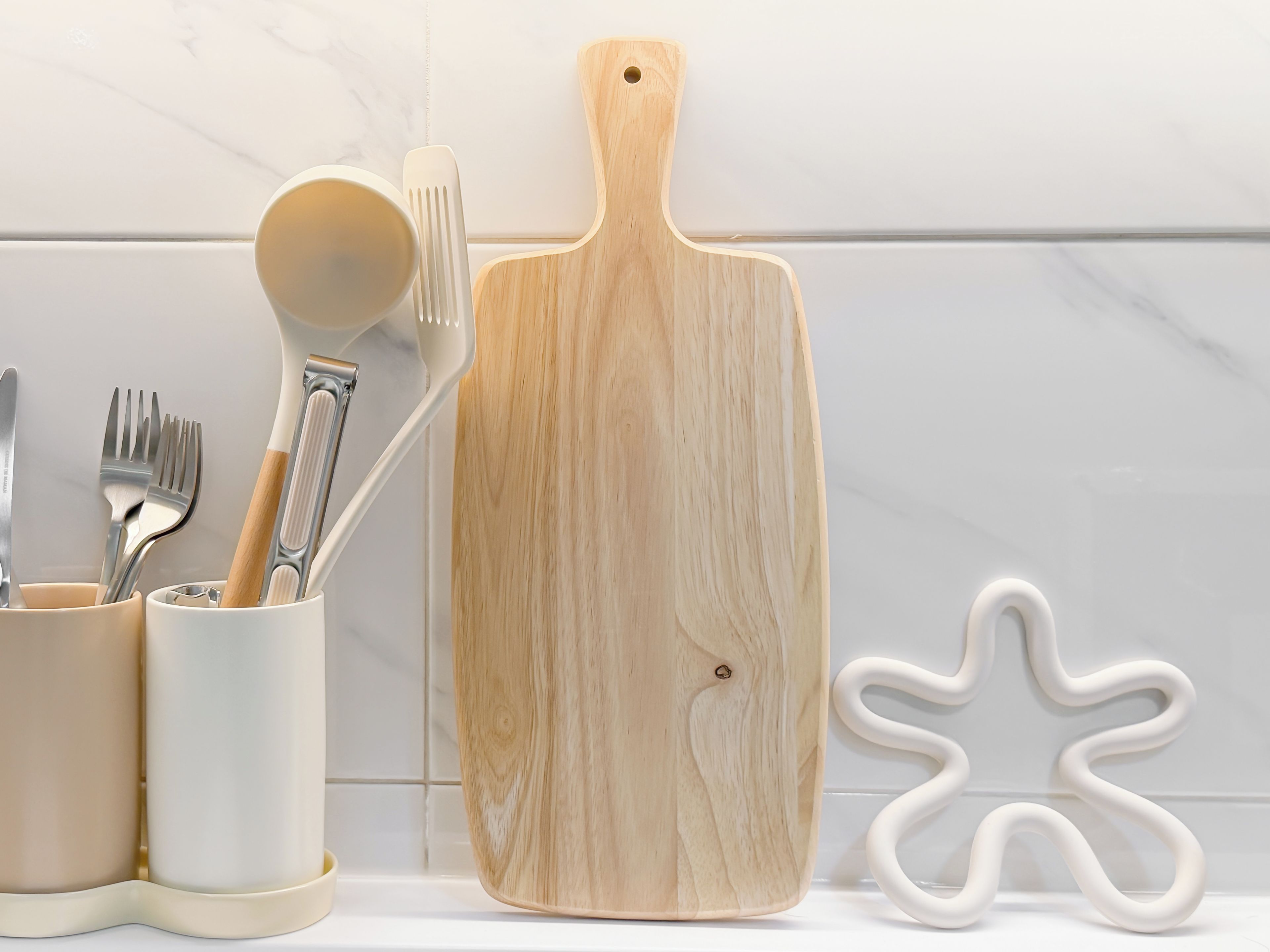 A kitchen counter with a wooden cutting board, a white ceramic spoon, and a white ceramic fork holder.
