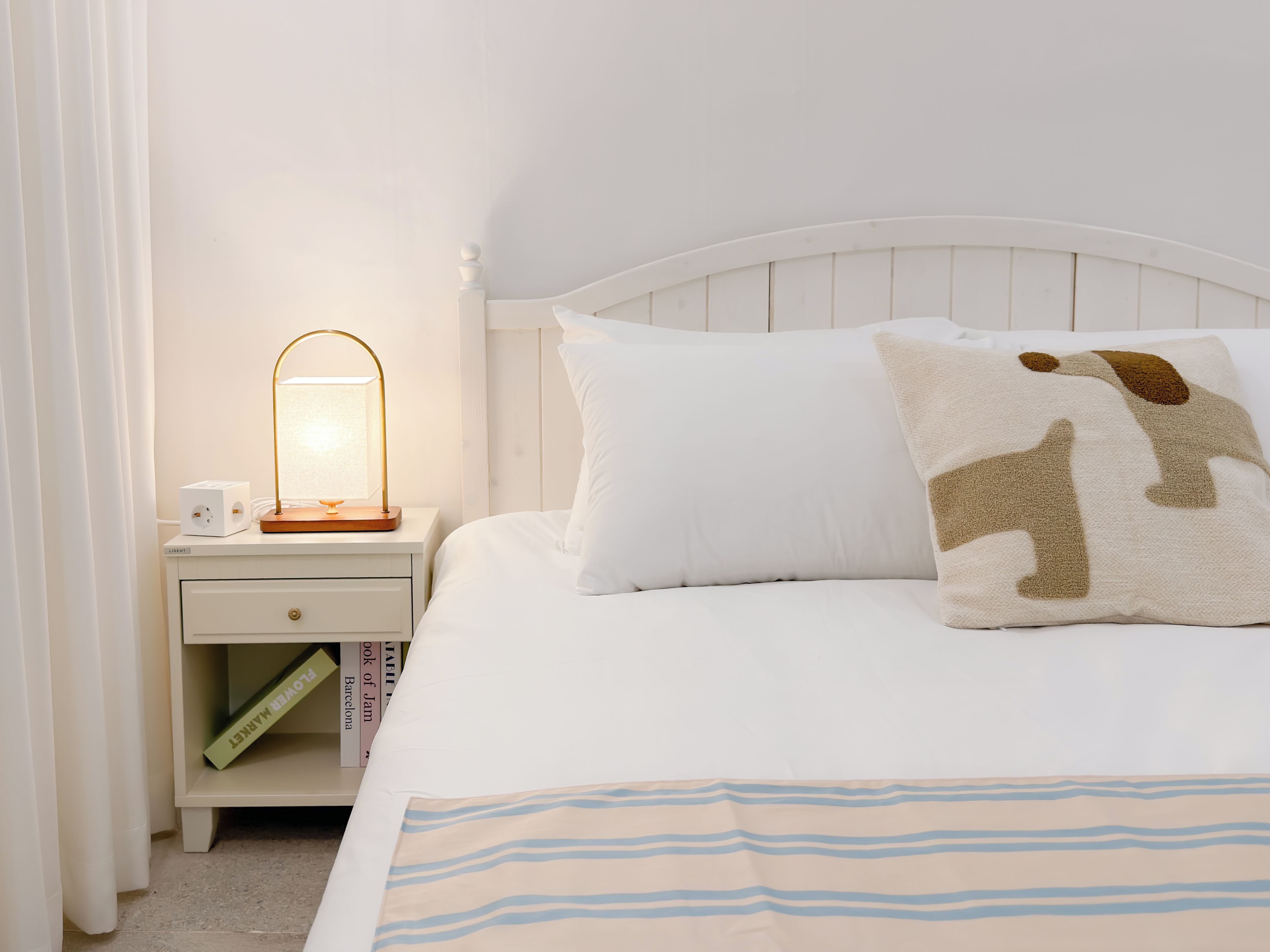 A neatly made bed with a striped blanket, a white nightstand with a lamp, and a book titled 'Book of Jam'.
