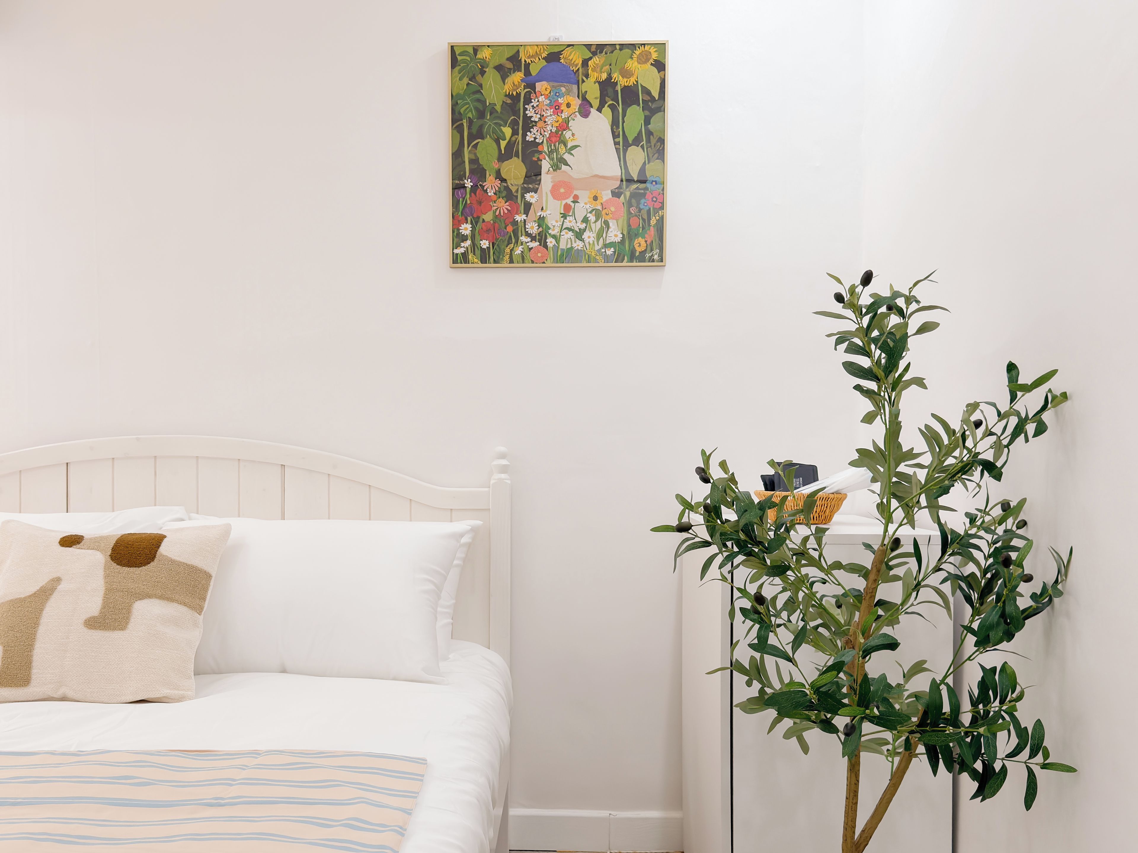 A bedroom with a white bed, a single pillow, a striped blanket, a wall-mounted painting, and a potted plant.