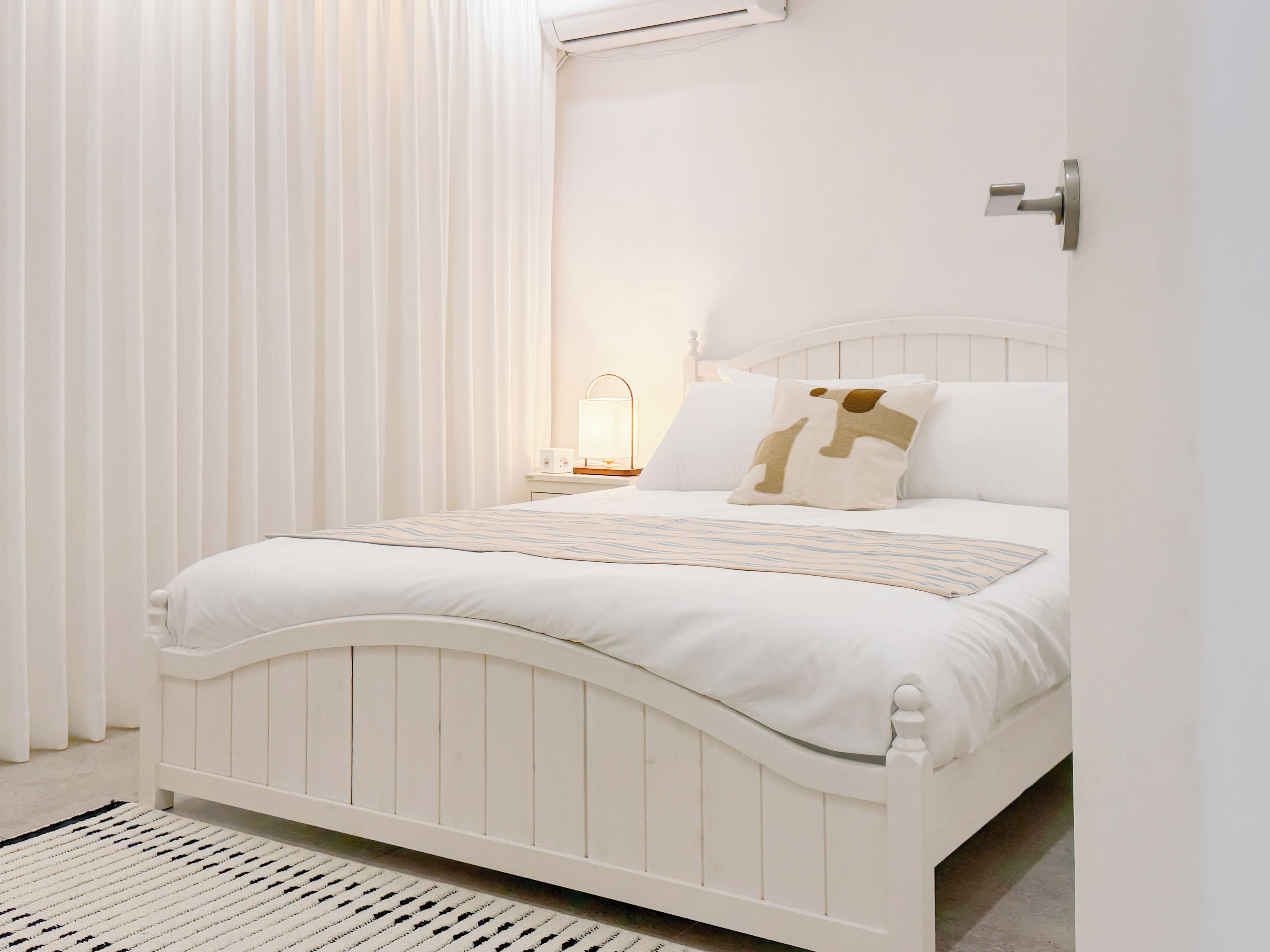 Ein weißes Bett mit Kopfteil, ein Nachttisch mit Lampe und ein Fenster mit Vorhängen.
