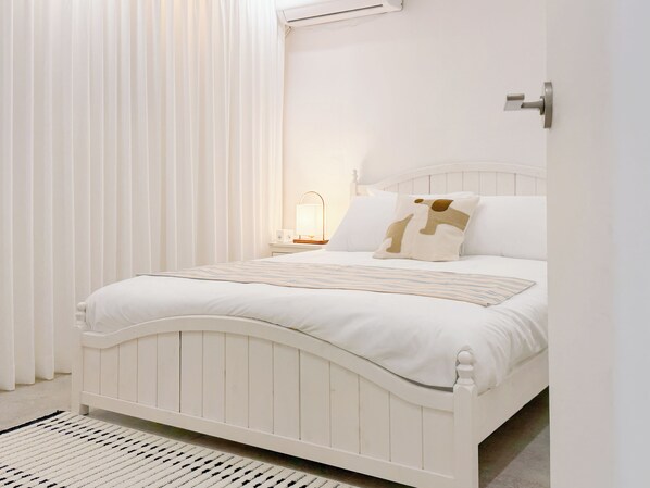 Una cama blanca con cabecera, una mesita de noche con lámpara y una ventana con cortinas.