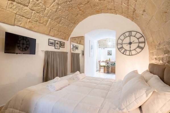 Un dormitorio con un arco de piedra, una cama con ropa de cama blanca, un televisor de pantalla plana y dos relojes de pared.