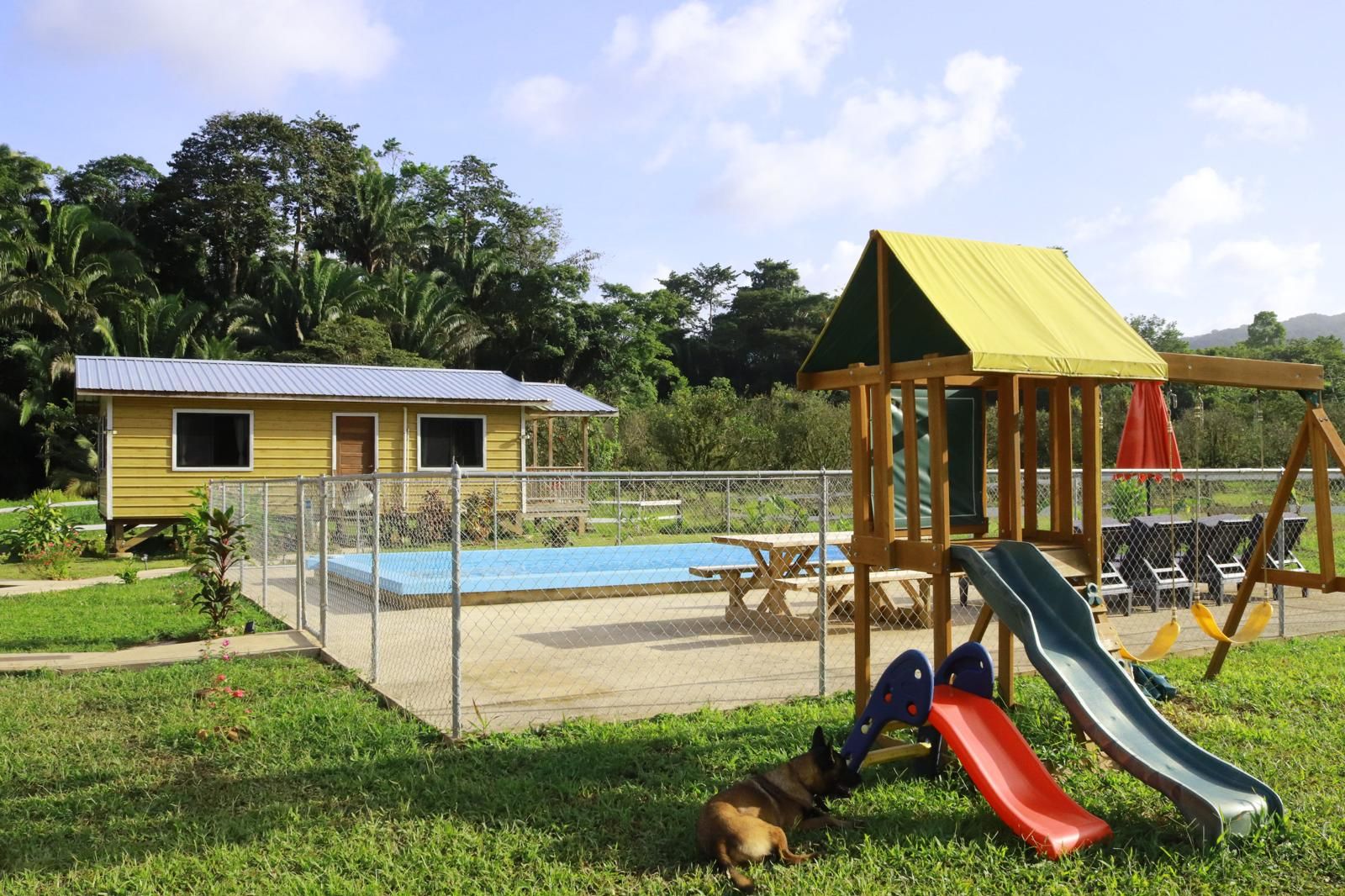 A fenced outdoor area with a playground, a small yellow building, and a pool.