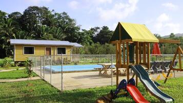 A fenced outdoor area with a playground, a small yellow building, and a pool.