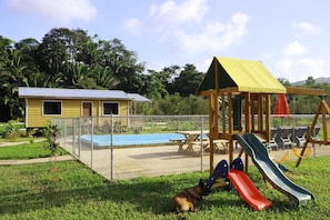 Un espacio exterior cercado con un parque infantil, un pequeño edificio amarillo y una piscina.