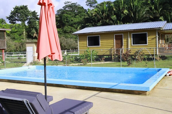 A swimming pool with a red umbrella, a yellow building with windows, and a fenced area with greenery.