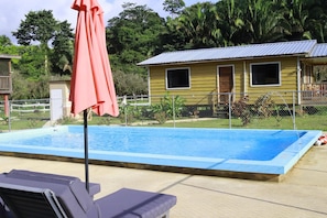 A swimming pool with a red umbrella, a yellow building with windows, and a fenced area with greenery.
