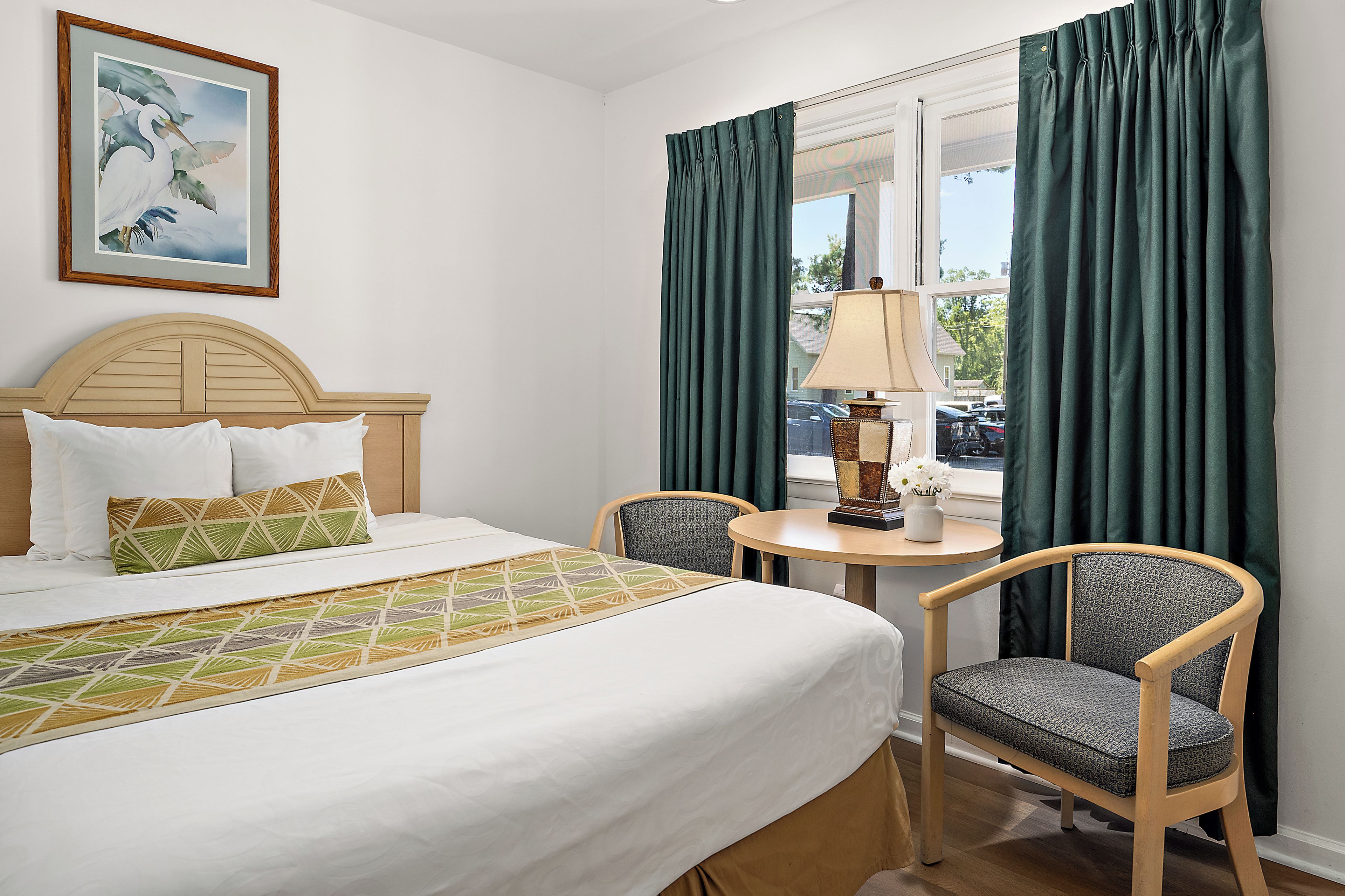 A hotel room with a bed, two chairs, a table with a lamp, a painting on the wall, and a window with curtains.