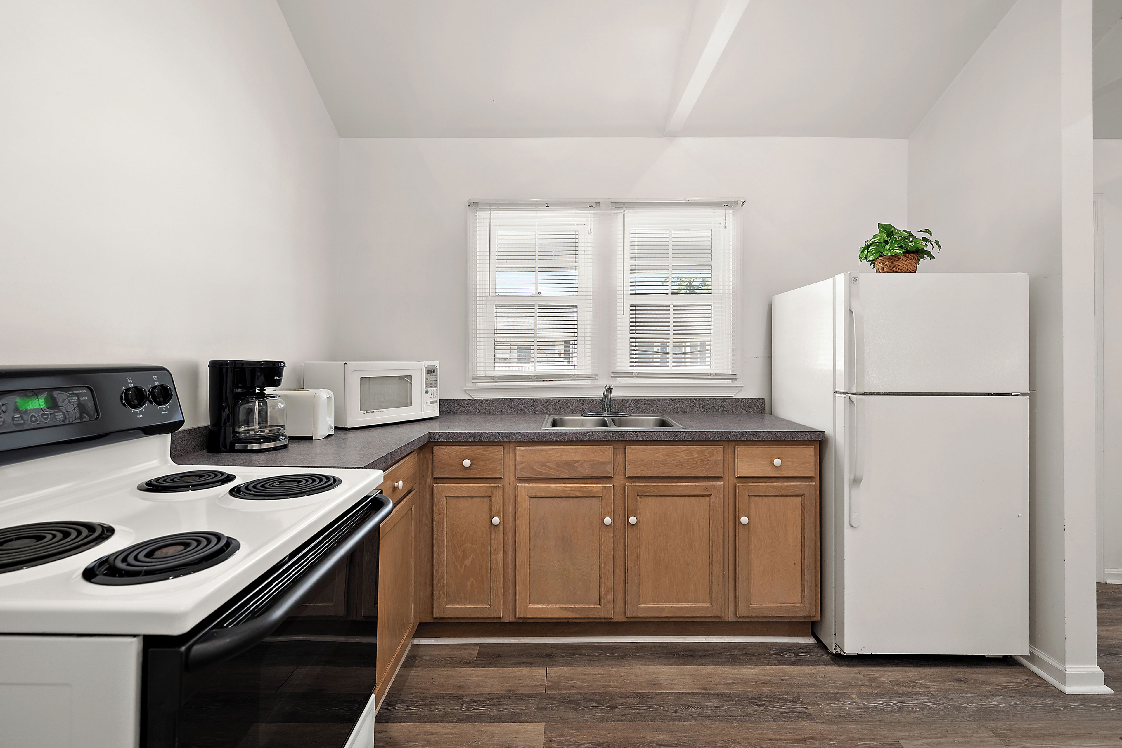 Una cocina con estufa blanca, microondas y refrigerador, gabinetes de madera, encimera de granito y una ventana con persianas.