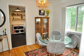 Un comedor con mesa de vidrio, dos sillas grises, un tapete estampado, un mueble de madera con puertas de cristal y una araña.