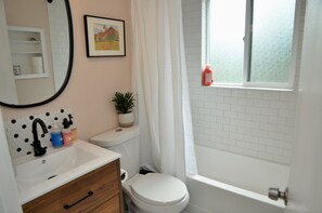 Un baño con lavamanos, inodoro y bañera blancos. Hay una ventana con vistas al exterior y una planta en maceta sobre el inodoro.