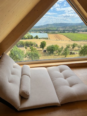Ein Zimmer mit großem Fenster und Blick auf eine ländliche Gegend, einer gepolsterten Bank und Holzbalken.