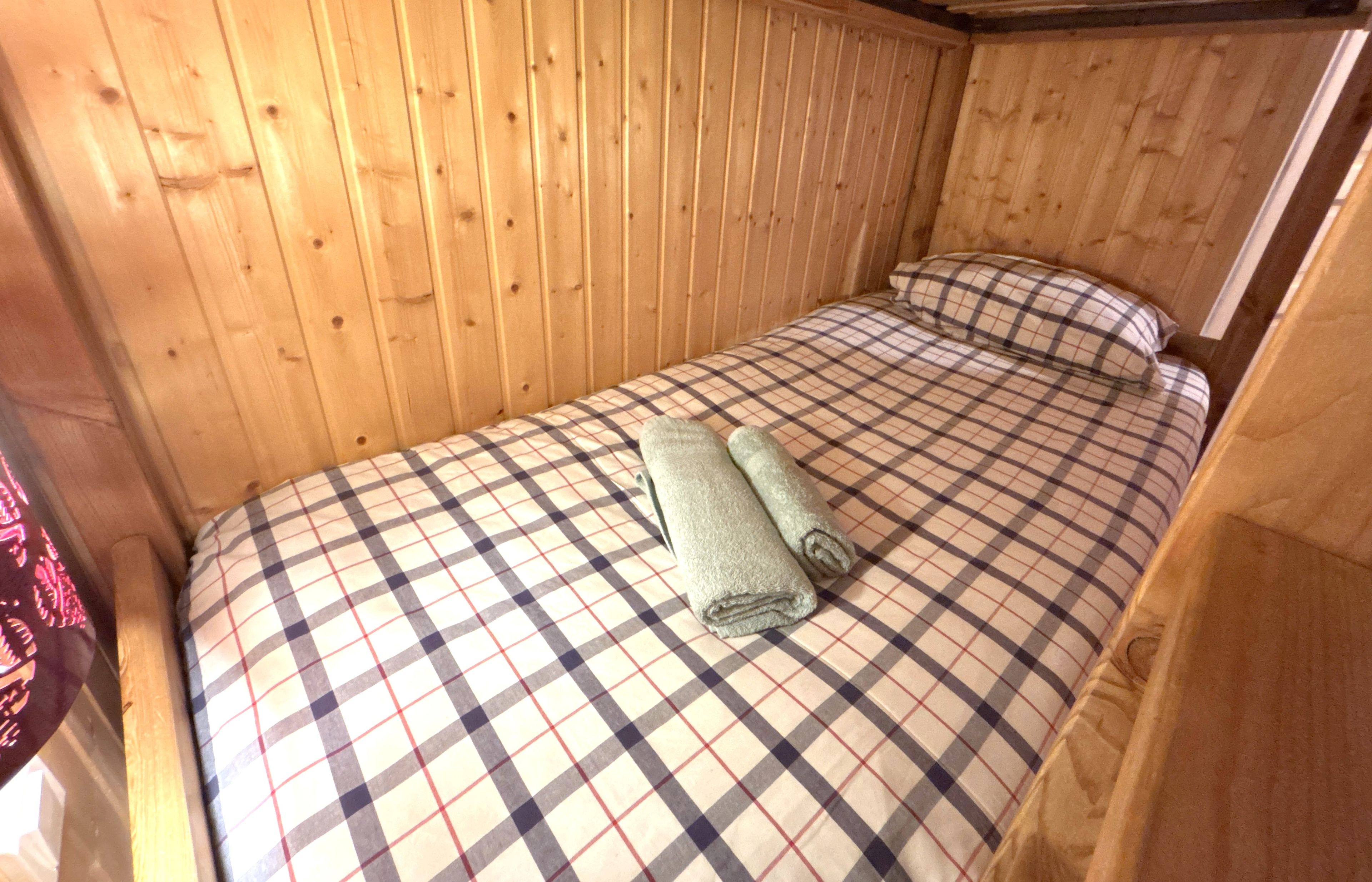 A single bed with a checkered bedspread and a rolled-up towel, wooden walls, and a headboard.