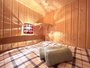 Ein Zimmer in einer Holzhütte mit einem Bett, einem Fenster mit einem roten, gemusterten Vorhang und einem Holz-Kopfschild.