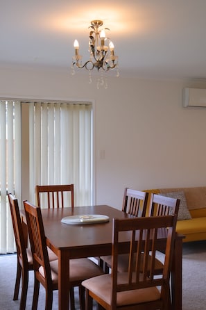 A dining area with a wooden table and chairs, a chandelier, and a sliding glass door with blinds. - Beauty near the Ocean spacious four-bedroom home (New Plymouth)