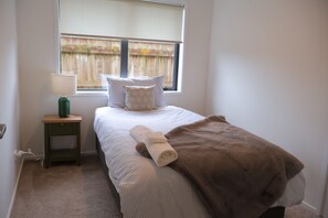 A bedroom with a bed, a nightstand, a lamp, a window with blinds, and a view of a wooden fence outside. - Beauty near the Ocean spacious four-bedroom home (New Plymouth)